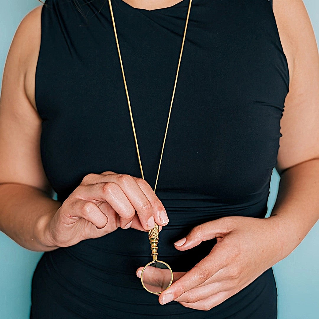 Person holding a gold necklace with a magnifying glass pendant against a blue background