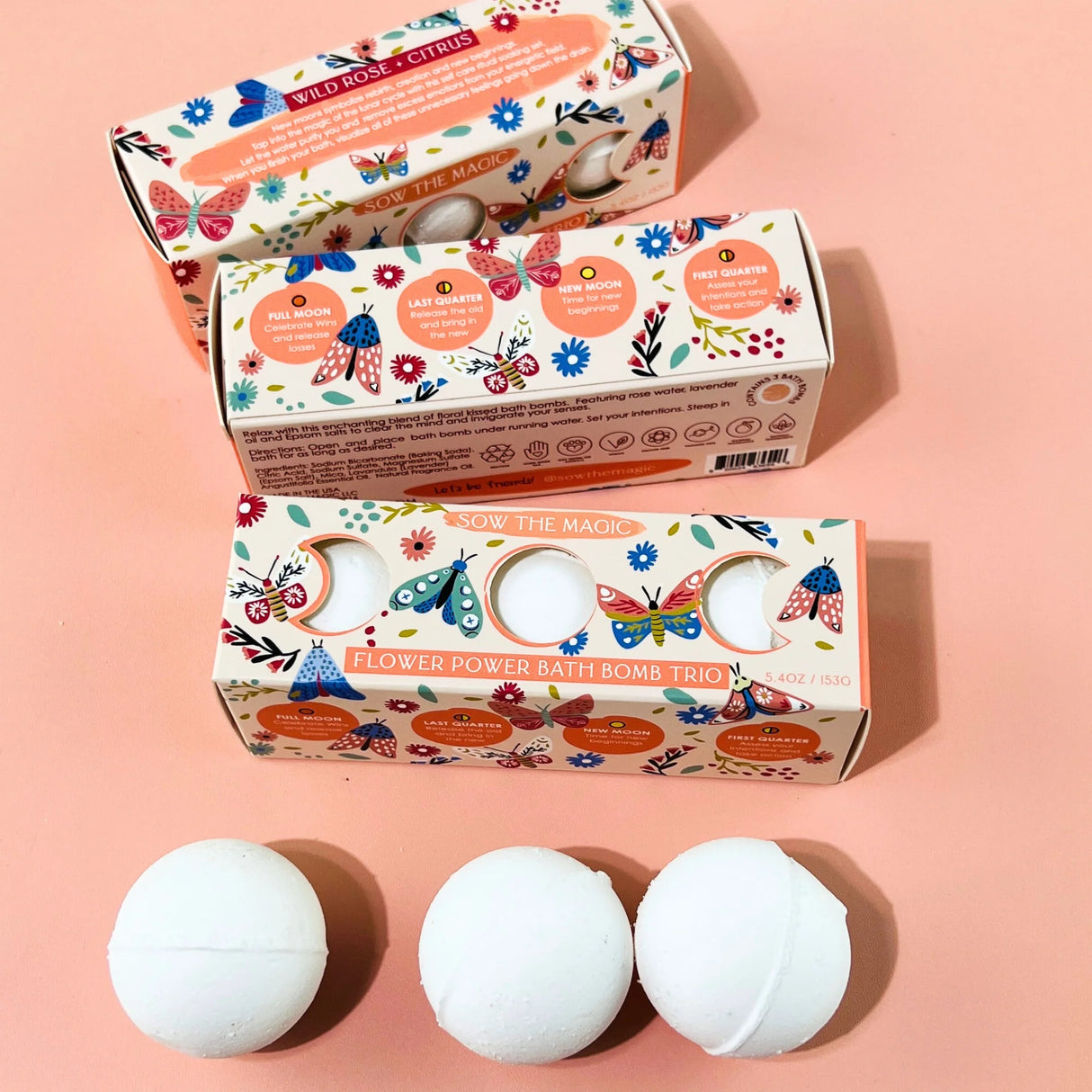 Three white bath bombs in front of their decorative boxes on a pink background