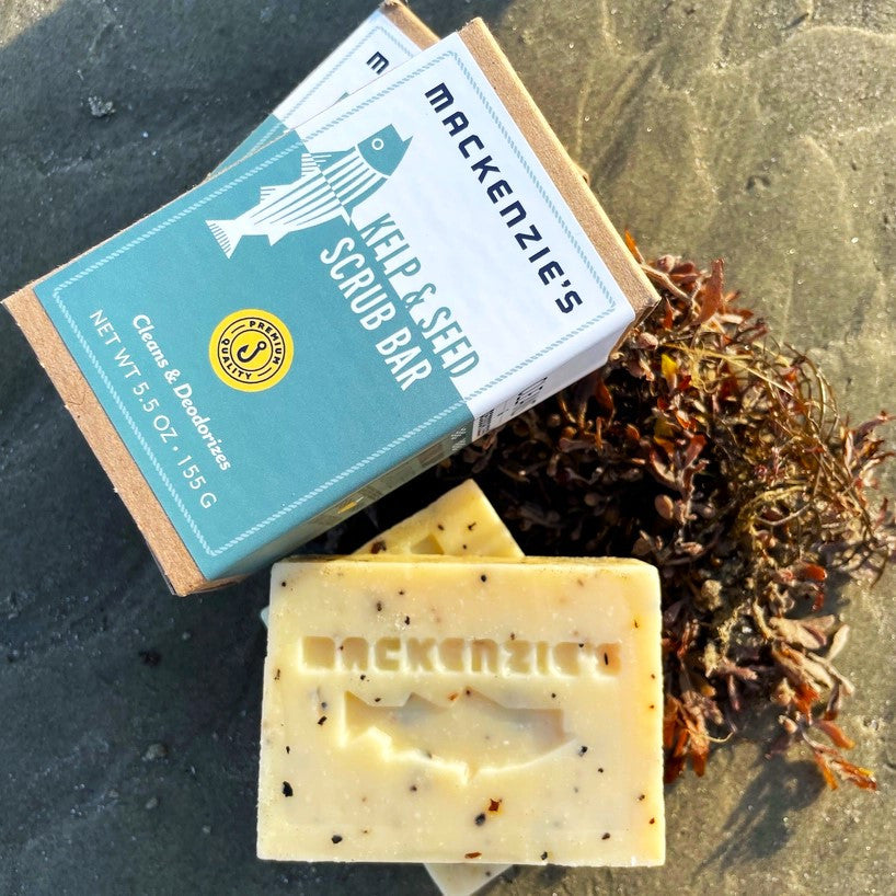 The Kelp & Sea Scrub bar out of the package showing the bits of kelp and seeds in the bar alongside the Kelp & Sea Scrub green and white packaging on a sandy beach with seaweed