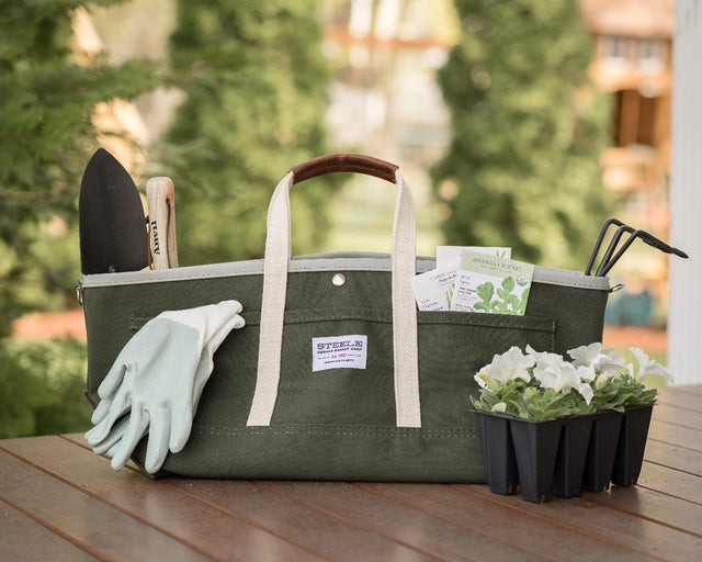 the garden tote loaded with tools, gloves, and seed packets sitting on a table alongside a pack of white flowers