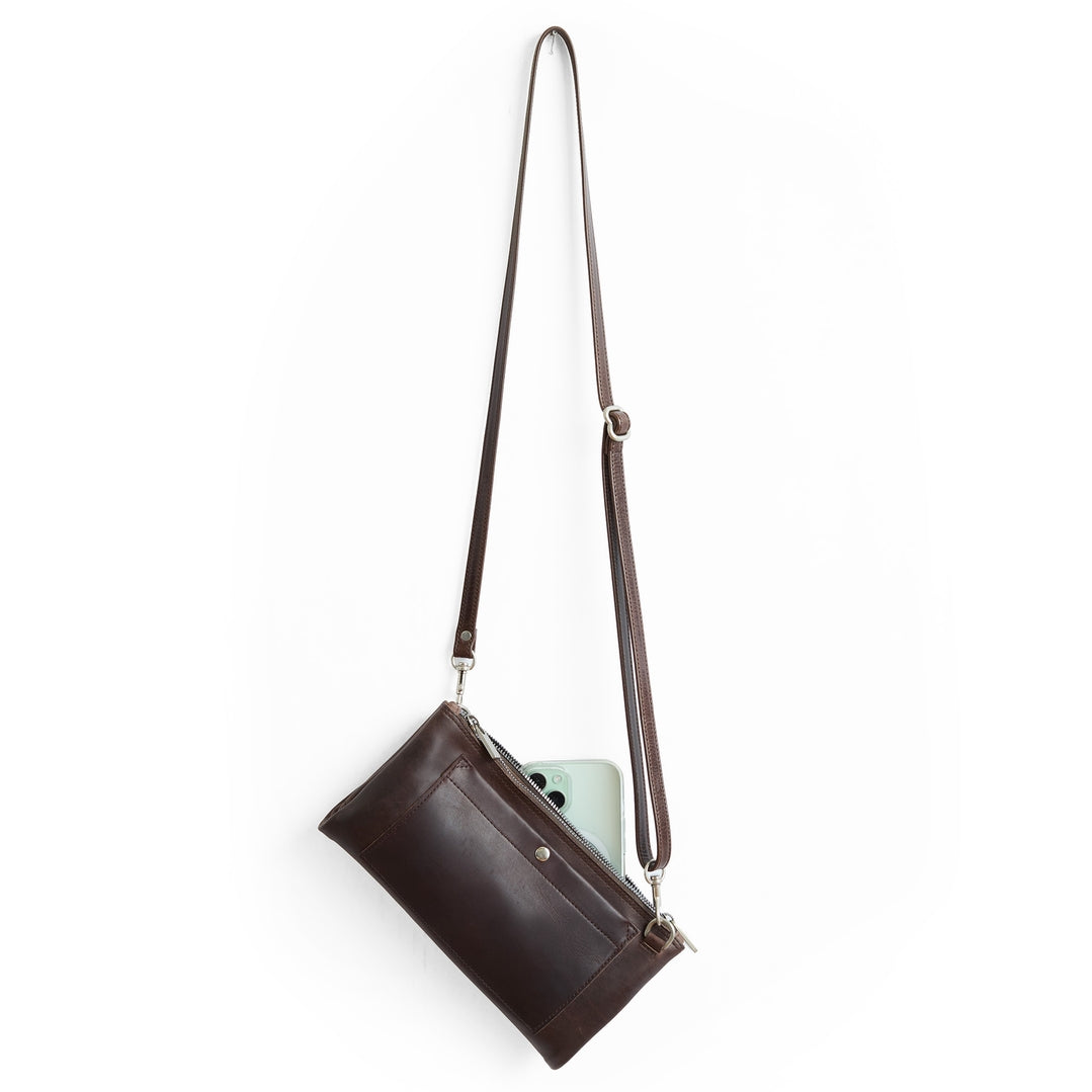 A dark brown leather crossbody bag with a long adjustable strap, shown with a light green smartphone tucked into its side pocket, against a solid white background.