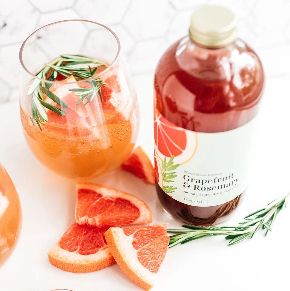 a bottle of the Grapefruit and Rosemary mixer next to a glass of prepared beverage that is garnished with sprigs of rosemary and slices of fresh grapefruit