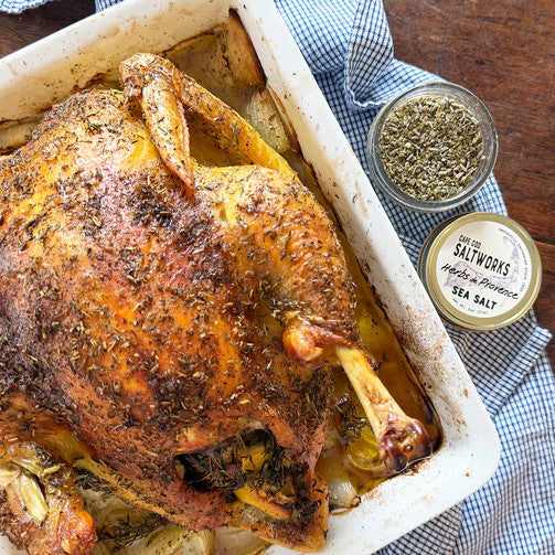 a large baking dish with a whole roasted chicken dusted with herbs alongside an open jar of herbs de provence sea salt