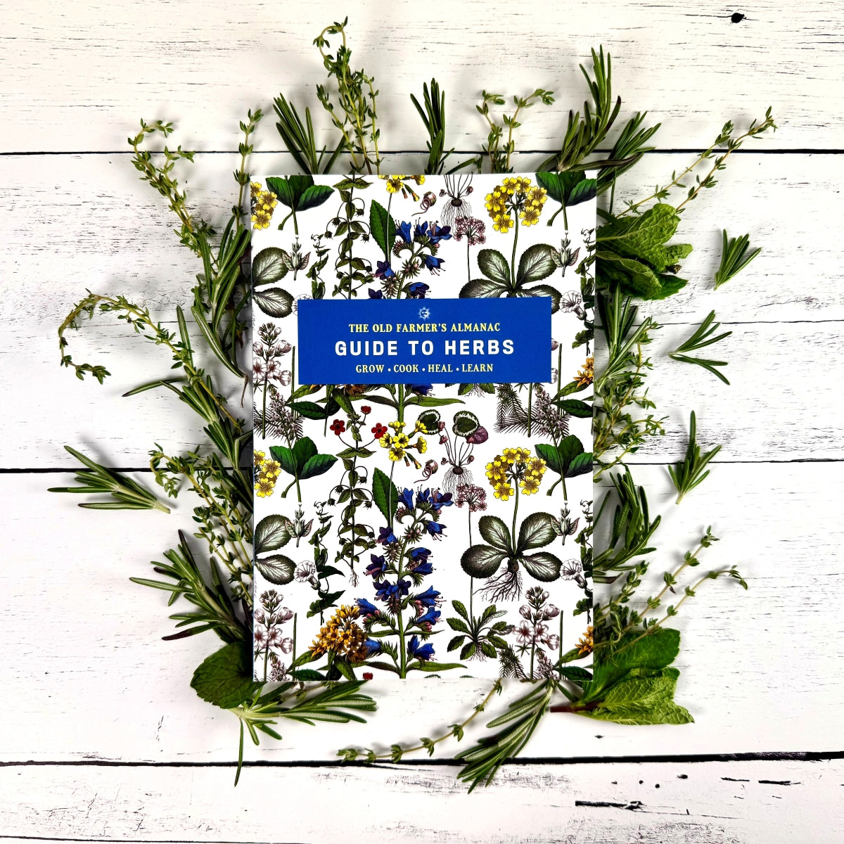 Book titled 'The Old Farmer's Almanac Guide to Herbs' surrounded by various herbs on a wooden surface.