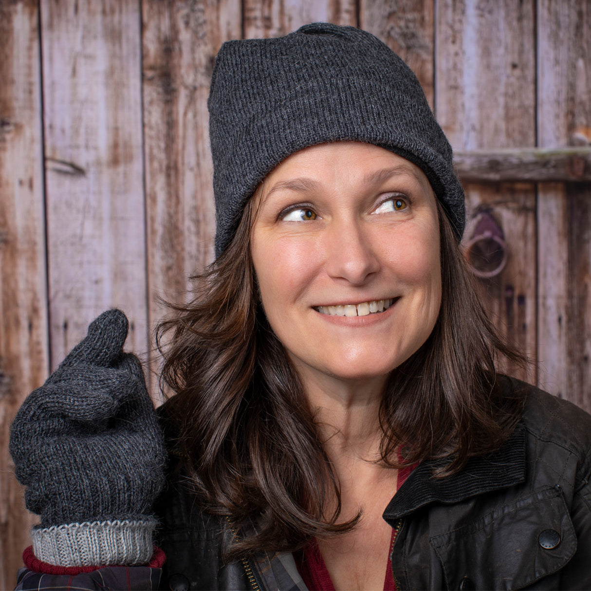a smiling woman stands in front of a rustic barn door and wears the gray iditarod hat and matching gloves