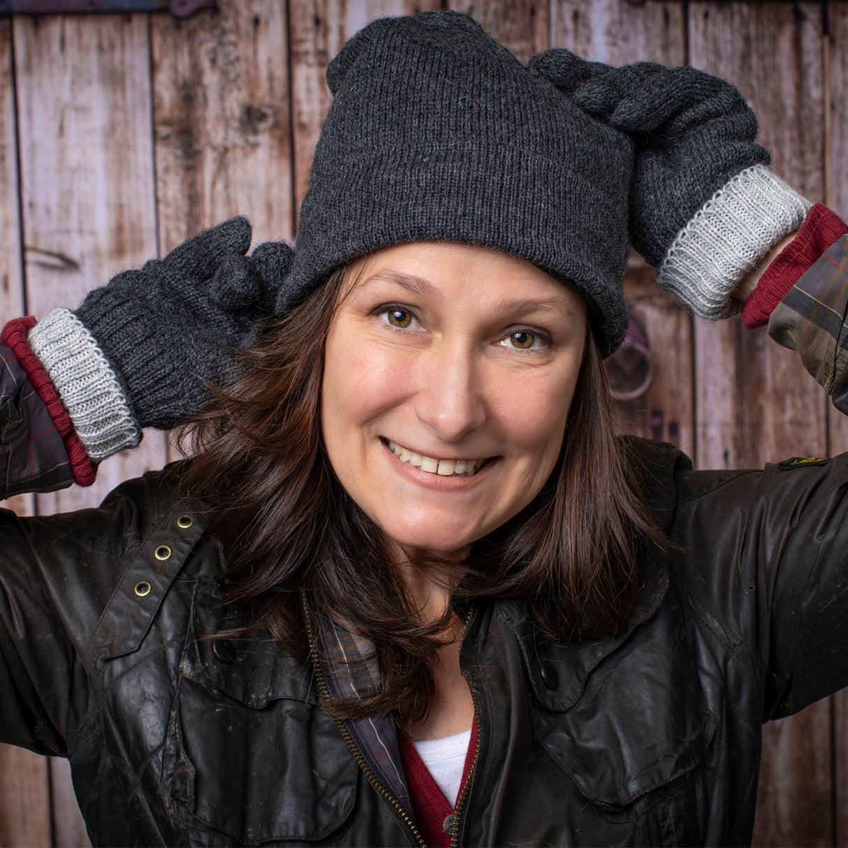 a smiling woman stands in front of a rustic barn door wearing the gray iditarod hat and matching gloves - she holds her hands behind her head playfully