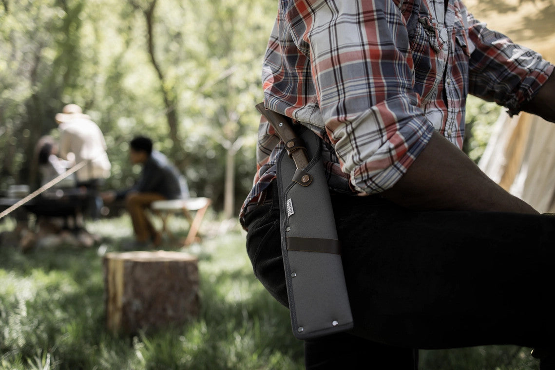 close up of the japanese nata hatchet in its sheath on attached to a man's belt as he stands at a lush forest campsite