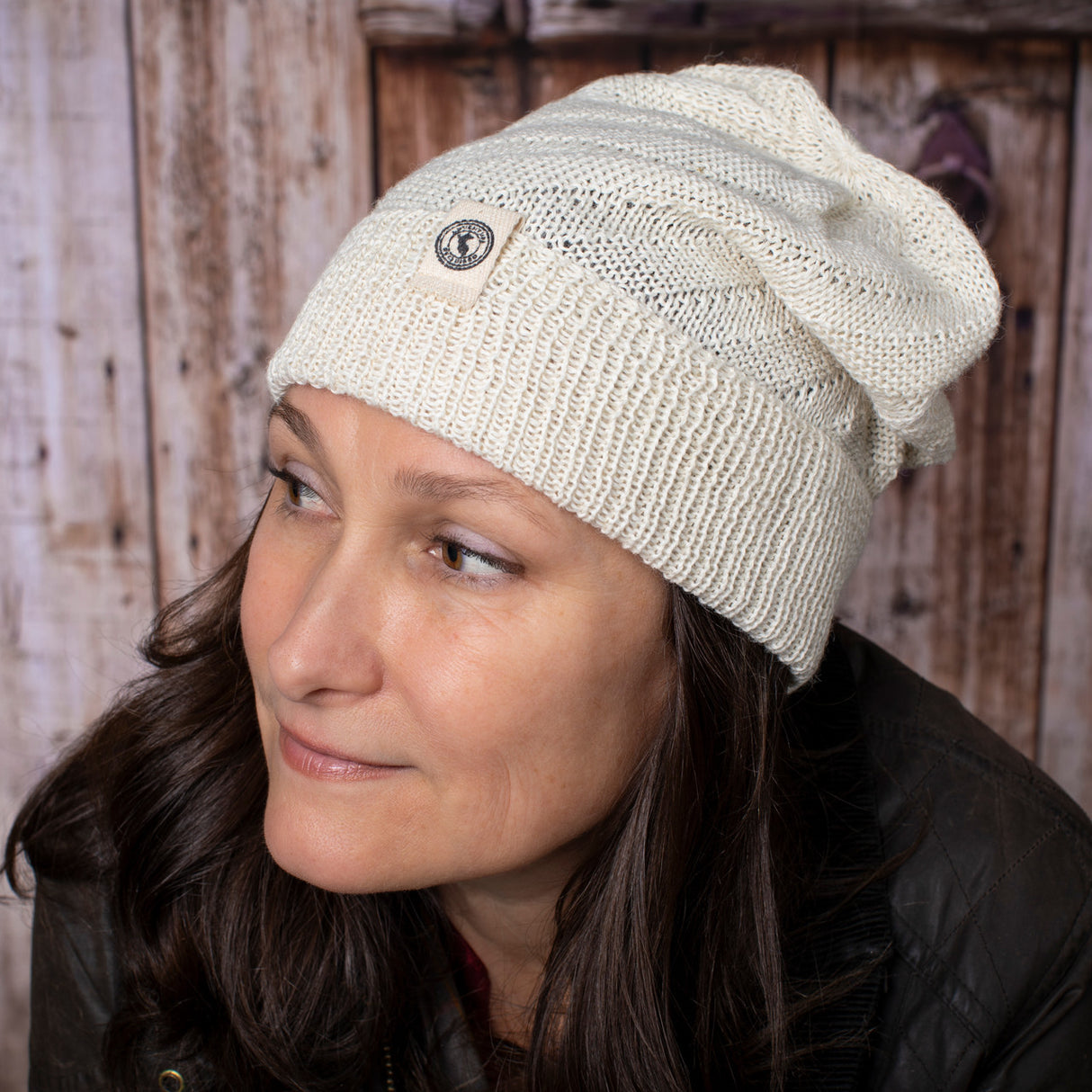 a fair skinned, brunette female model wearing the Cream beanie - she is smiling softly and posed in front of a rustic barn door