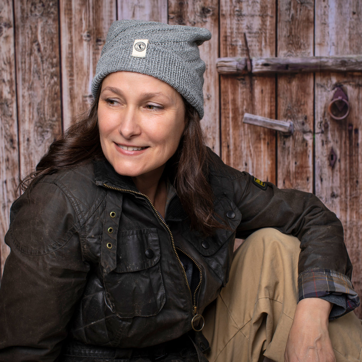 a fair skinned, brunette female model wearing the Kingsley beanie in Gray. She is smiling and posing casually with her arm resting on her knee and sitting in front of a rustic barn door