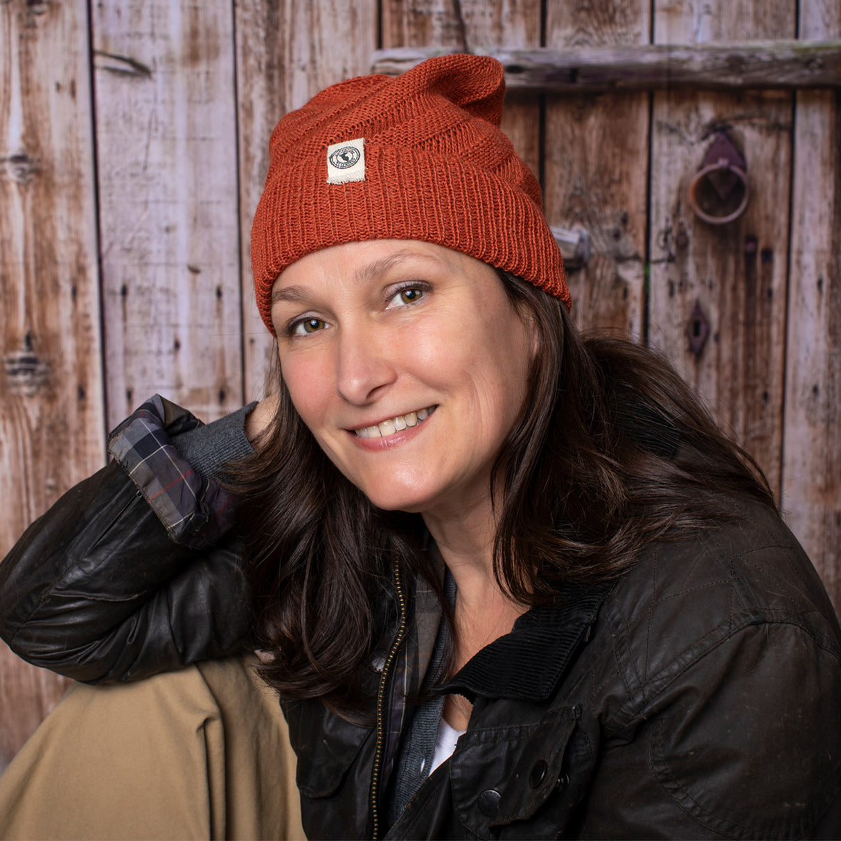 a fair skinned, brunette female model wearing the Kingsley beanie in Rust. She is smiling and posing casually with her arm resting on her knee and sitting in front of a rustic barn door