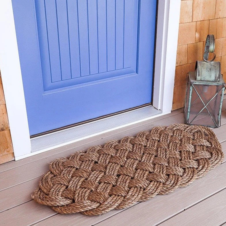 a side angle view of a large manila rope doormat with an intricate hand woven pattern on a doorstep in front of a sky blue door