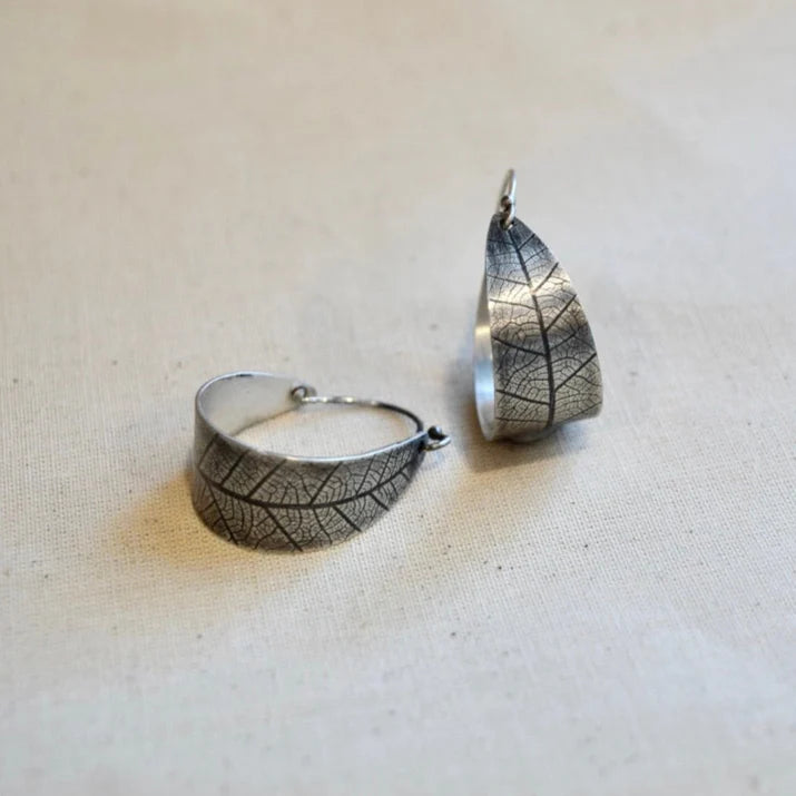 Silver leaf-patterned hoop earring and pendant on a beige fabric background