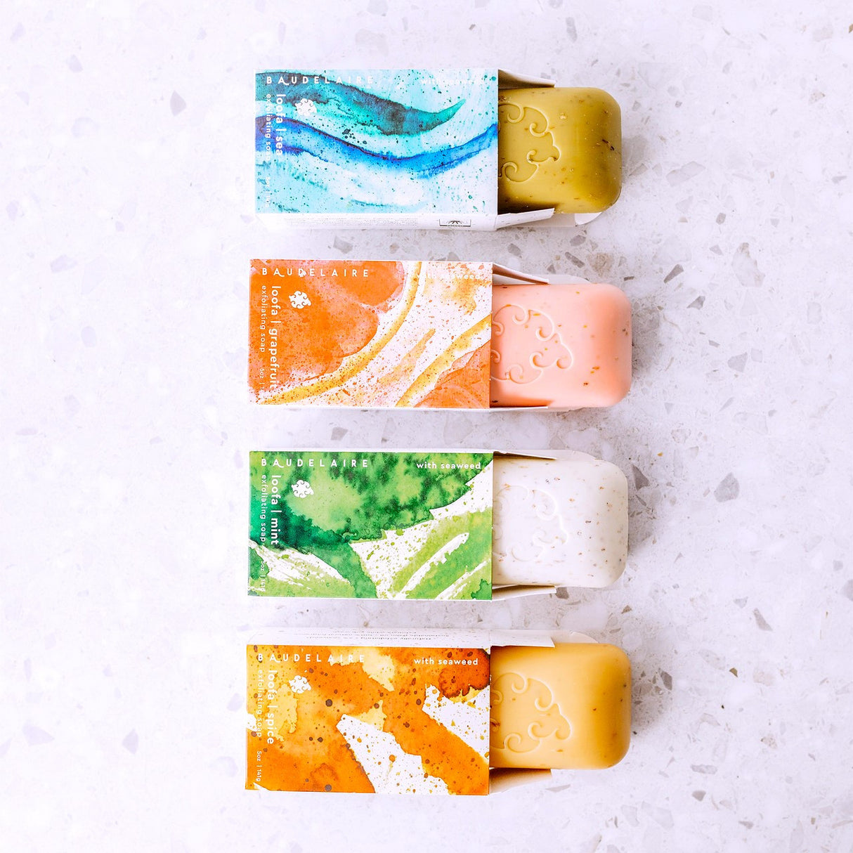 all four scents of the Loofa soaps in a vertical line with each soap peeking out of its package showing both the box design and soap color