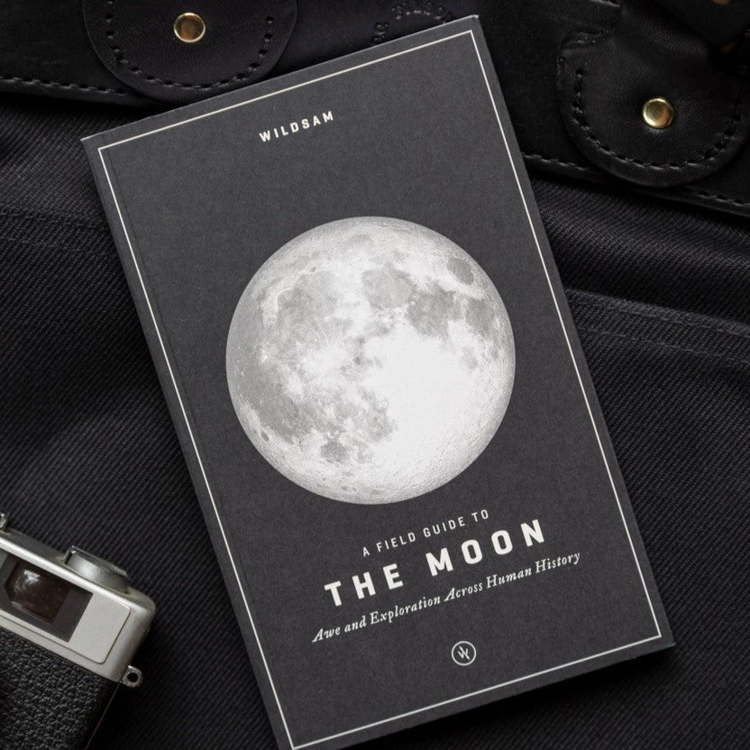 the moon guide resting on a black leather bag alongside a camera showing that this field guide is sized for travel