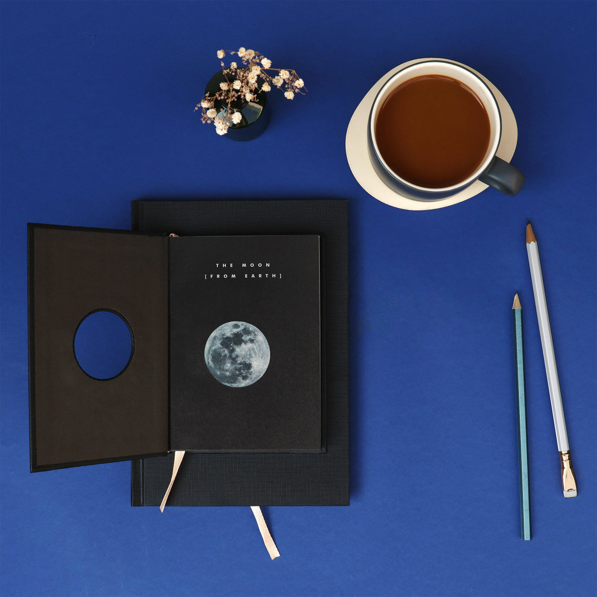 Black notebook with moon illustration on a blue background, accompanied by a cup of tea and pencils.