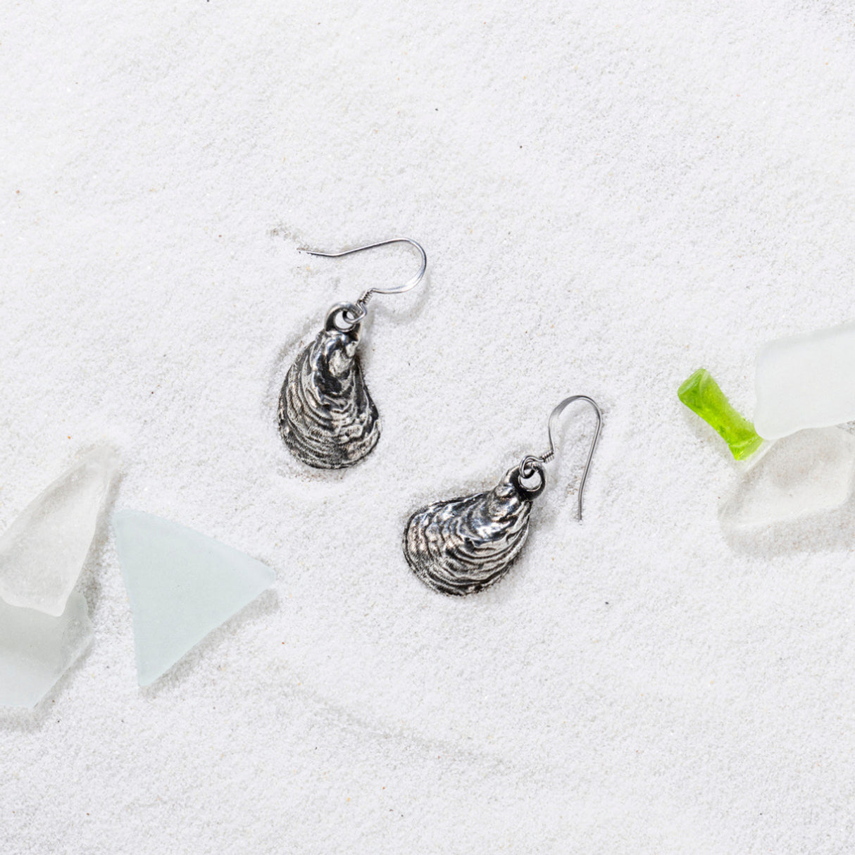 a pair of pewter oyster shell shaped earrings on a white sandy beach surrounded by bits of sea glass giving a coastal atmosphere