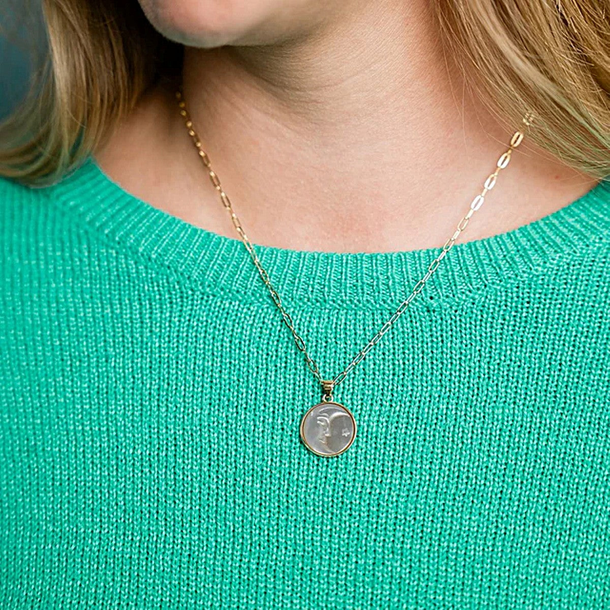 Person wearing a gold necklace with a round pendant on a teal sweater