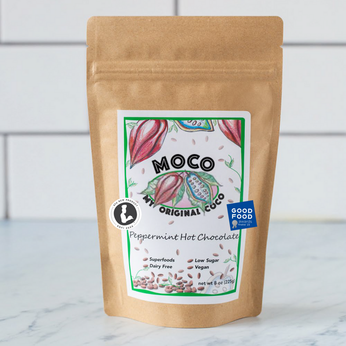 a kraft hot cocoa package with a white label with red and green illustrations of cocoa beans that reads "MOCO My Original Coco - Peppermint Hot Chocolate" on a marble counter with white subway tile background