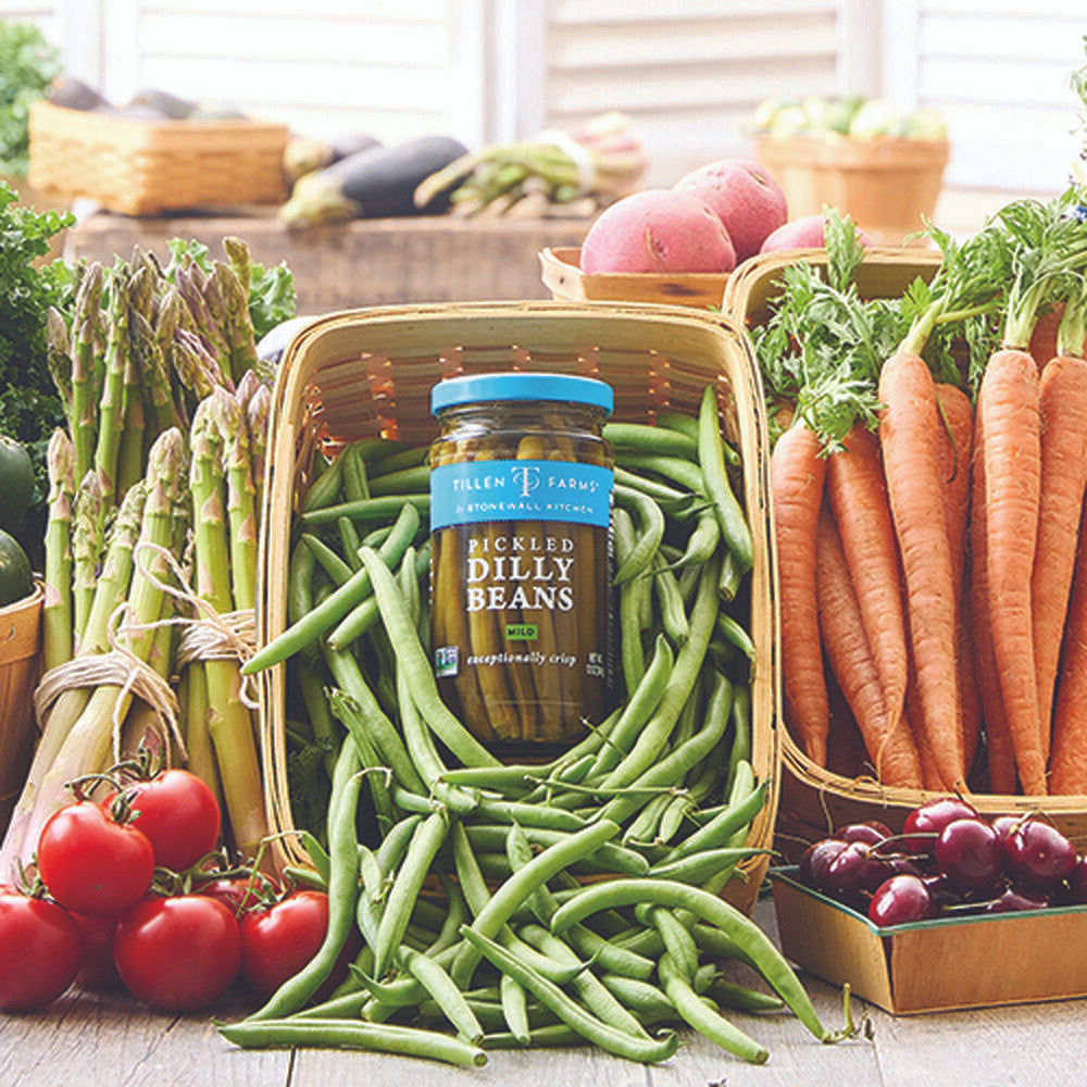 a jar of dilly beans nestled in an overflowing pile of fresh green beans, surrounded by a beautiful fresh veggie spread of asparagus, tomatoes, carrots, and more