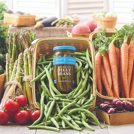 a jar of dilly beans nestled in an overflowing pile of fresh green beans, surrounded by a beautiful fresh veggie spread of asparagus, tomatoes, carrots, and more