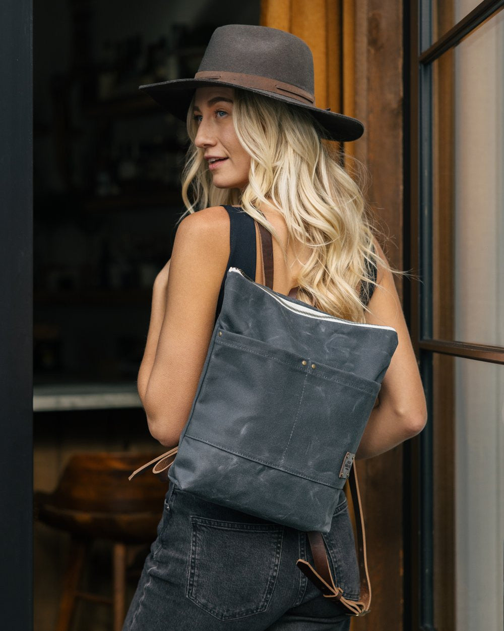 a blonde woman with a wide brim hat stands in a doorway with the Day Pack in Ash slung over her left shoulder