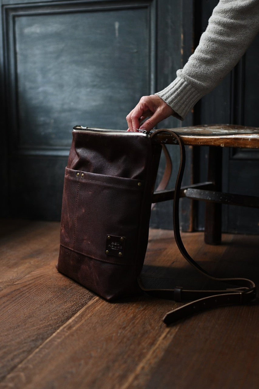 the day pack in chestnut rests against a chair while a hand gently reaches into the top of the pack