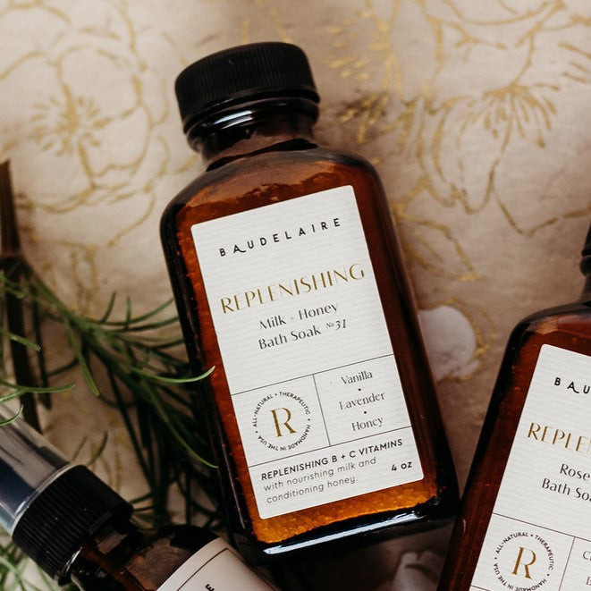an amber colored apothecary style bottle with a black cap and off white label that reads "Baudelaire - REPLENISHING- Milk + Honey Bath Soak no. 31 - Vanilla - Lavender - Honey"