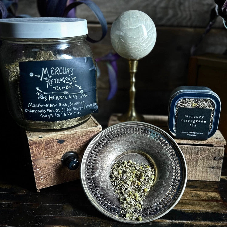 a table with a spread of tea in various containers - a large clear jar with a white lid and handwritten black label, a silver bowl tipping forward with loose leaf tea spilling out, and the silver tea tin