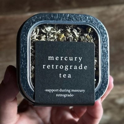 a close up of the mercury retrograde tea tin with elegant black label and clear top to show the loose leaf tea inside