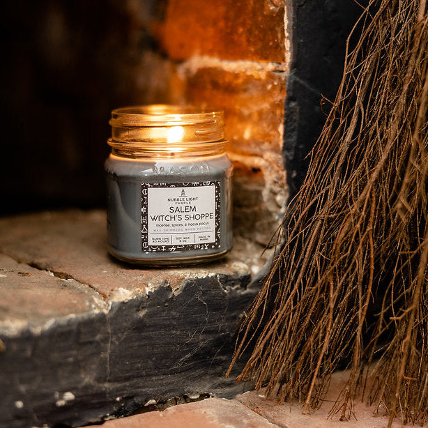 Candle labeled 'Salem Witches Shoppe' next to a broom in front of a fireplace.
