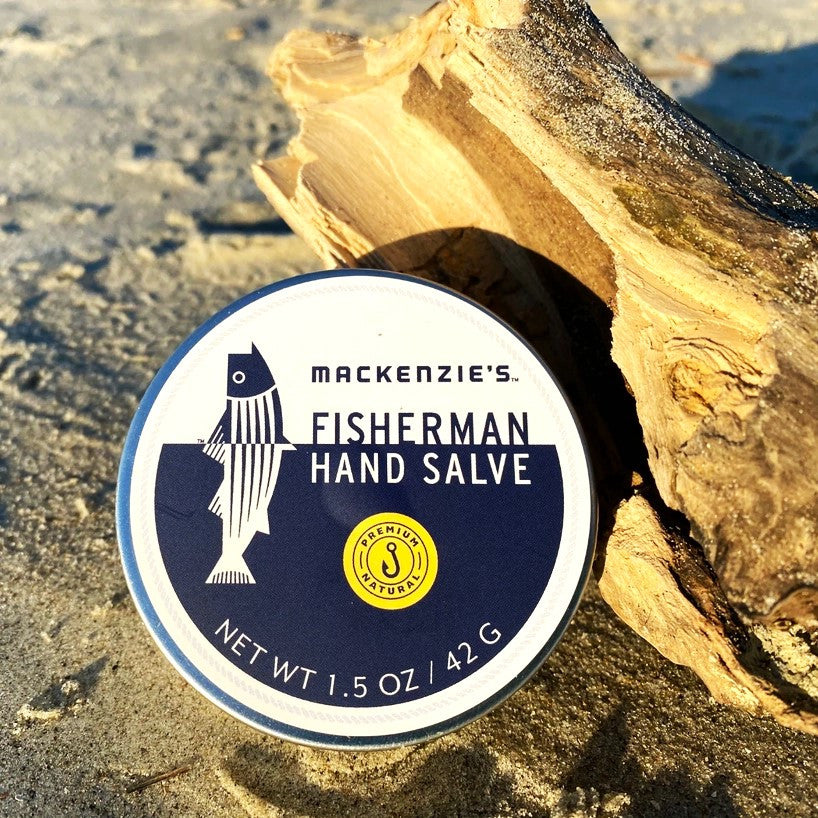 a tin of Mackenzie's Hand Salve on a sandy beach leaning up against a piece of driftwood