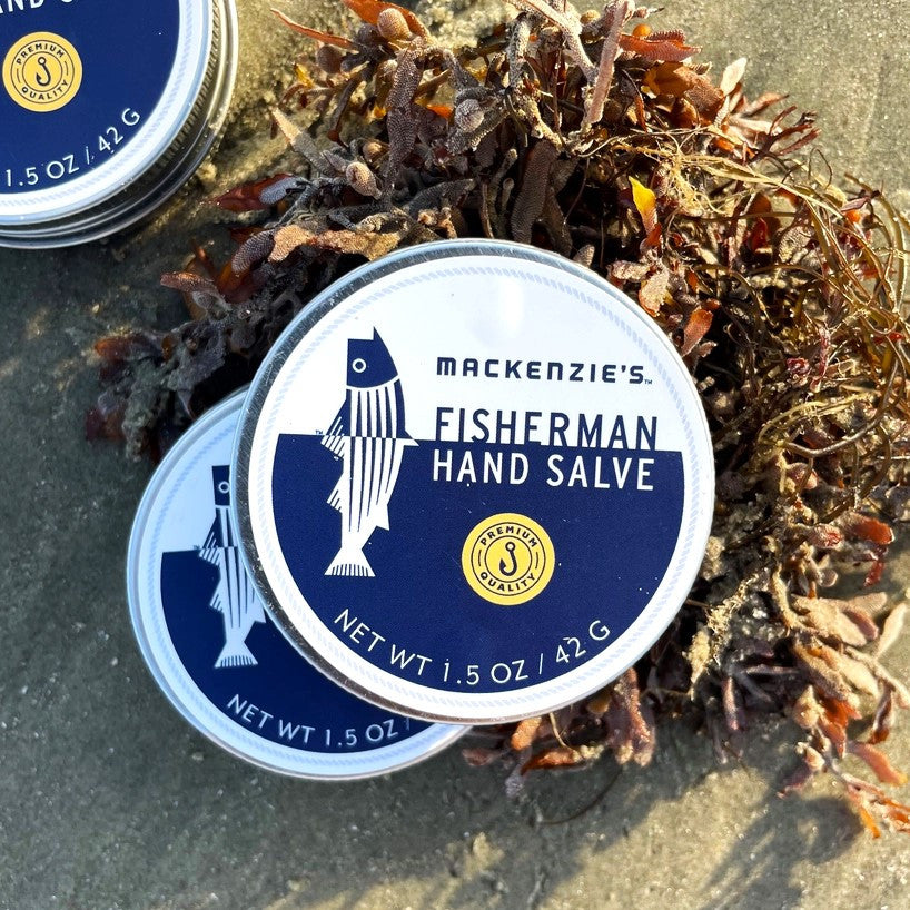 two tins of Mackenzie's Hand Salve on a sandy beach leaning up against a pile of seaweed