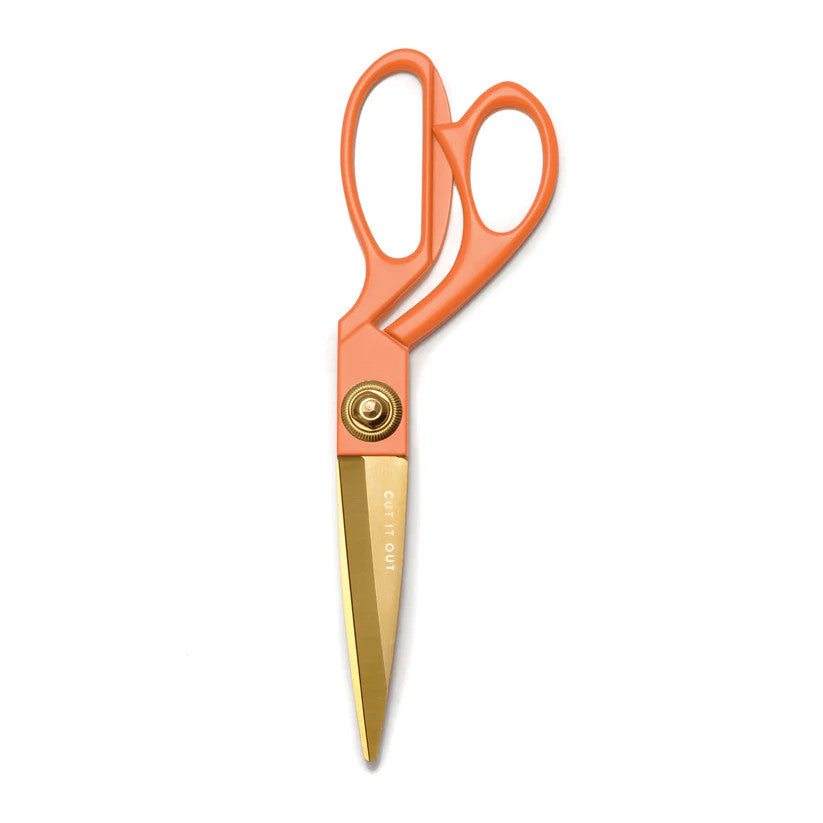 a vintage inspired pair of scissors with light orange enamel handles and gold blades etched with the phrase "Cut it Out" against a white background