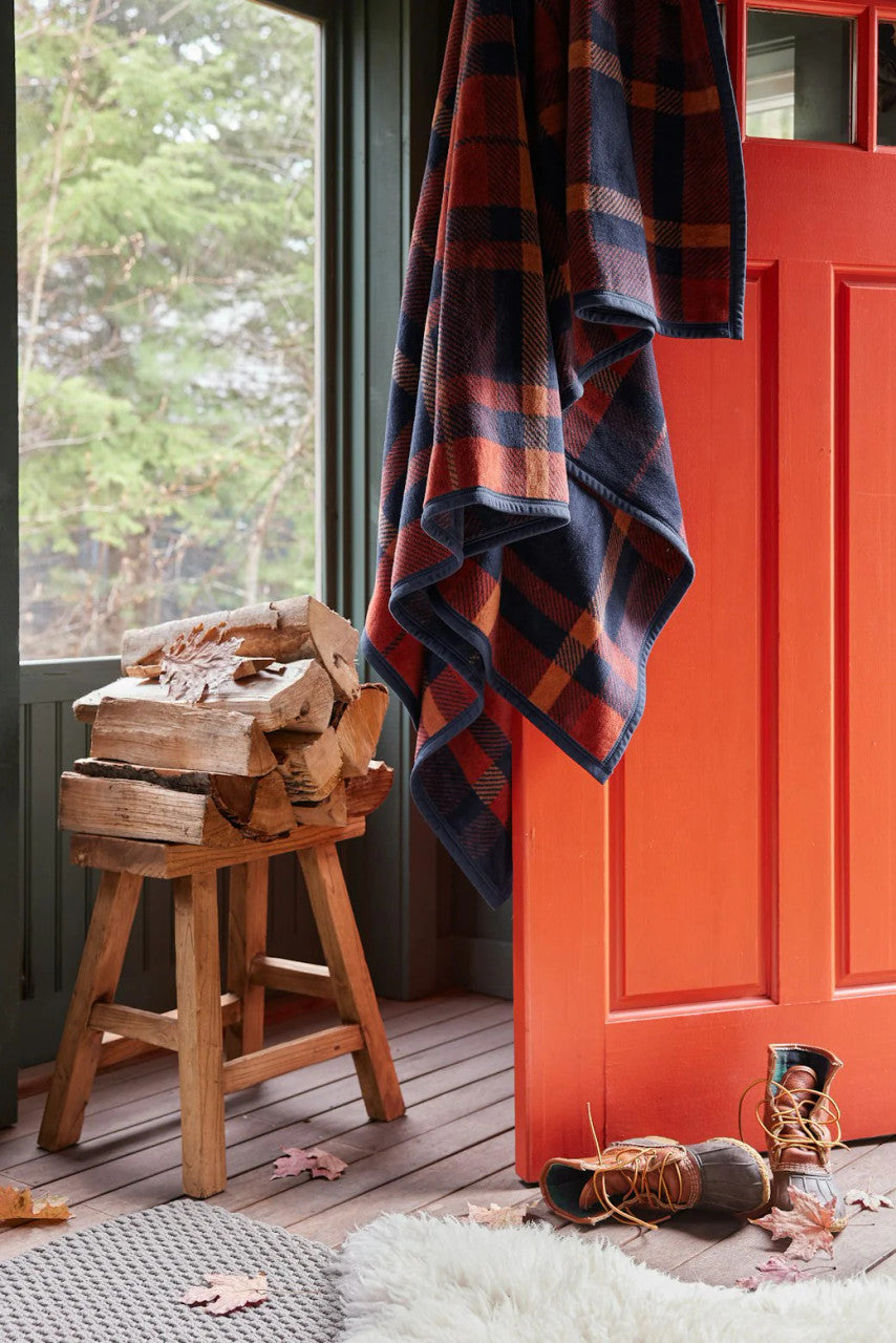 a rustic cabin scene with the sebago tartan blanket draped over an open, orange door with a pair of boots, chopped firewood, and a shearling rug nearby setting the mood for this cozy autumnal blanket
