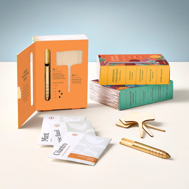 Collection of seed library packages, a gold dibbler tool, and small packets on a light surface with a light blue background