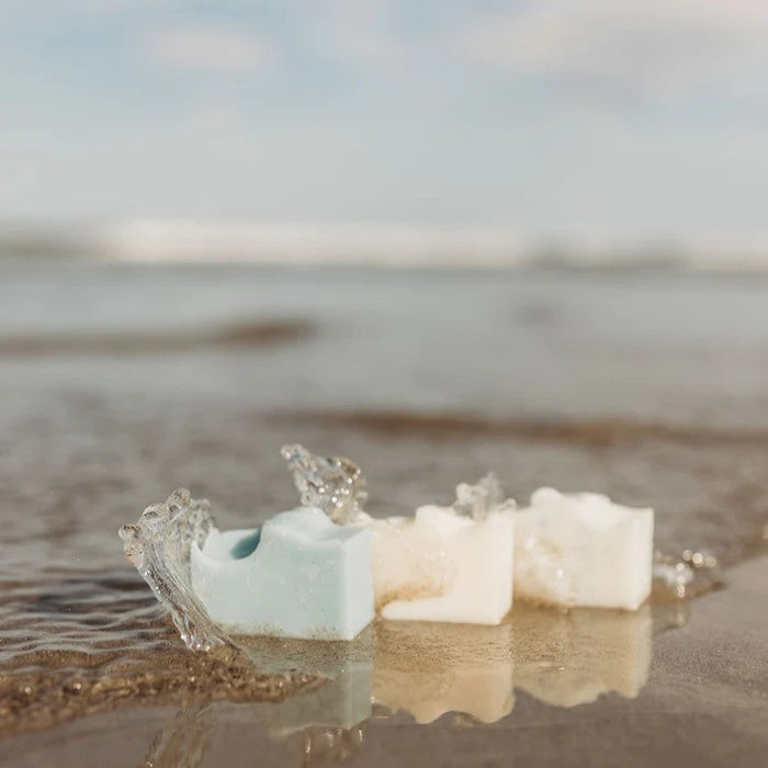 the sel de mer soap trio on the seashore with a small wave splashing against the bars of soap