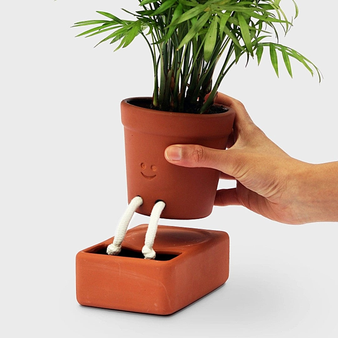Hand holding a terracotta pot with a plant, attached to a matching base with a smiley face design and cotton "legs" that drape into the watering reservoir at the base of the planter.