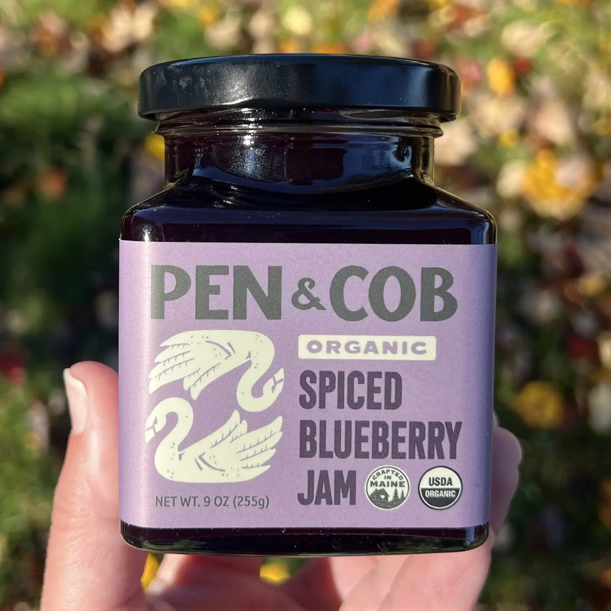 Jar of Pen & Cob Organic Spiced Blueberry Jam held in a hand with a blurred natural background
