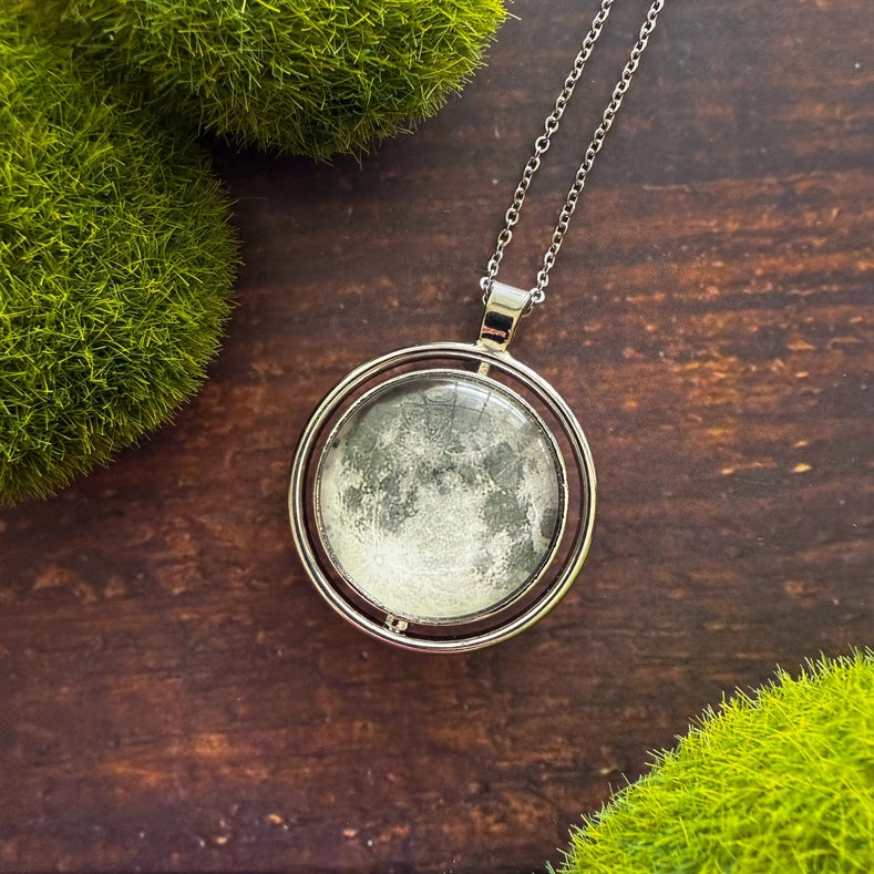 Moon-phase locket necklace on a wooden surface with green moss