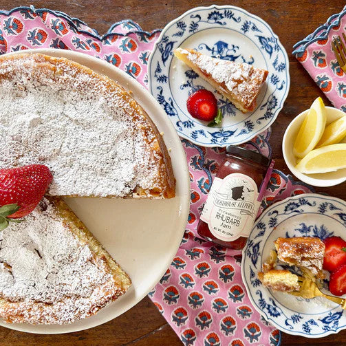 a jar of Strawberry Rhubarb jam on a table next to plated slices of cake garnished with strawberries and a large cake dusted with powdered sugar and garnished with a whole strawberry
