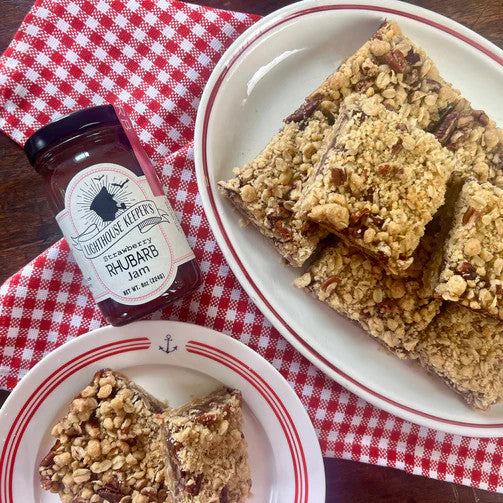 a jar of strawberry rhubarb jam on a red and white checkered cloth alongside a plate of oat squares