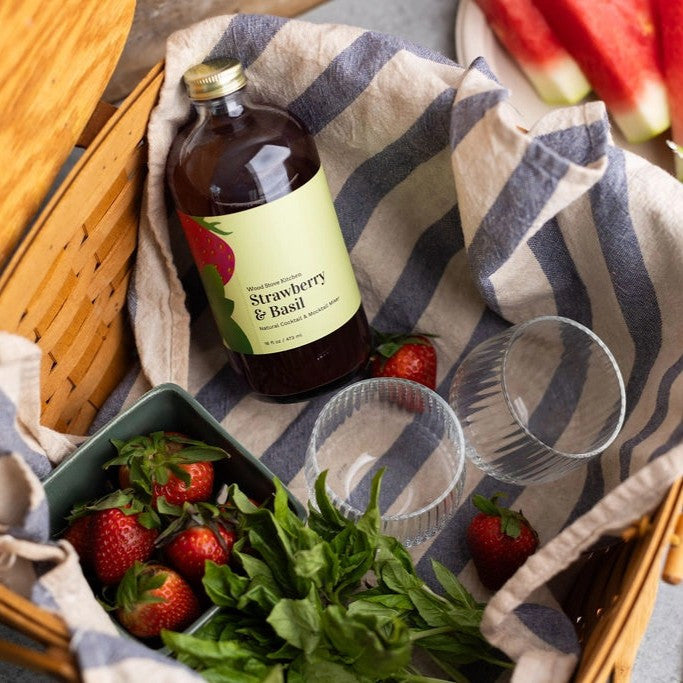 interior view of a picnic basket with a bottle of Strawberry & Basil mixer, two glasses, and fresh strawberries and basil leaves