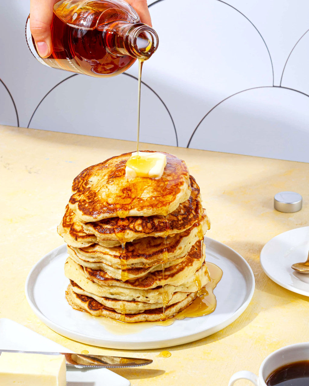 a bright kitchen table with a tall stack of pancakes on a plate with maple syrup being poured over them
