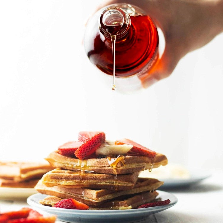 a hand holdling a bottle of maple syrup and pouring it onto a tall stack of waffles topped with strawberries