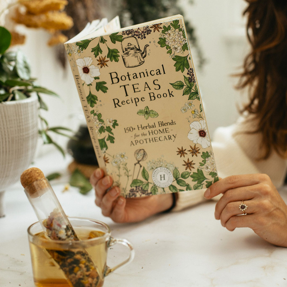 a person reading the botanical teas recipe book at a white table alongside a clear glass tea cup