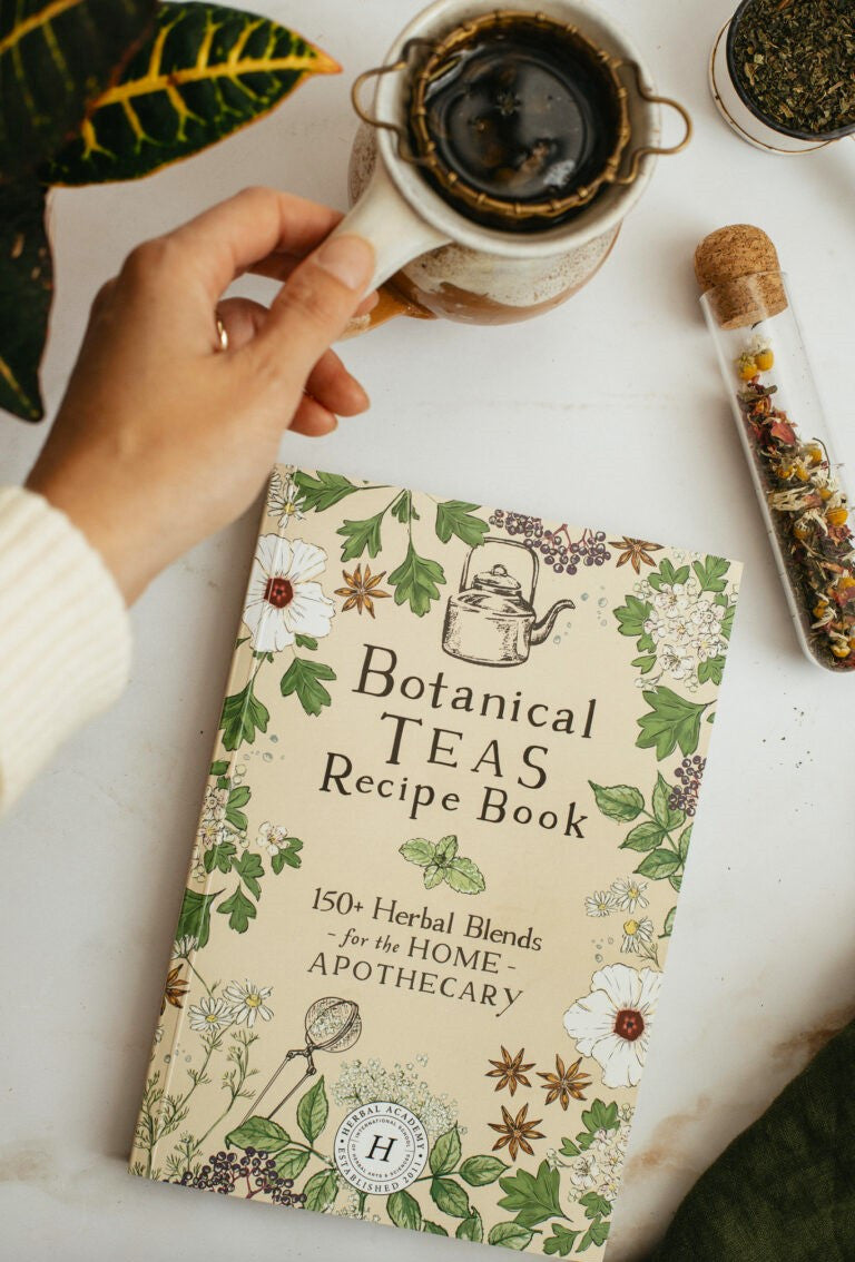 The botanical teas recipe book on a white table next to a vial of dried herbs next to a tea cup with strainer