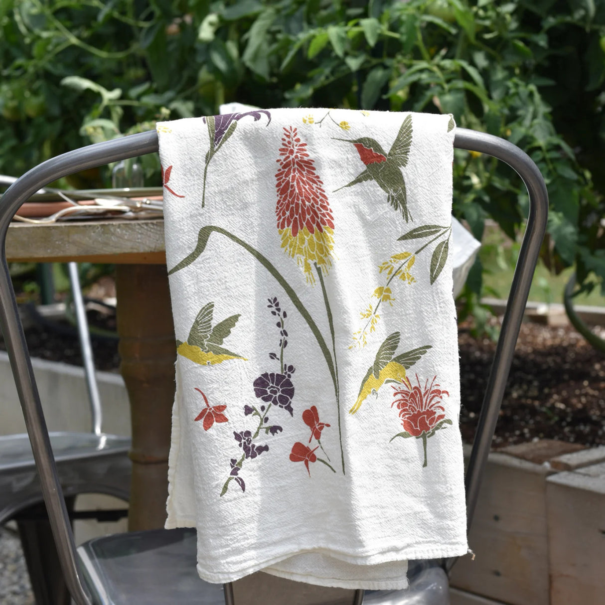 the hummingbird garden kitchen tea towel folded and layed over the back of a chair on a lush garden patio