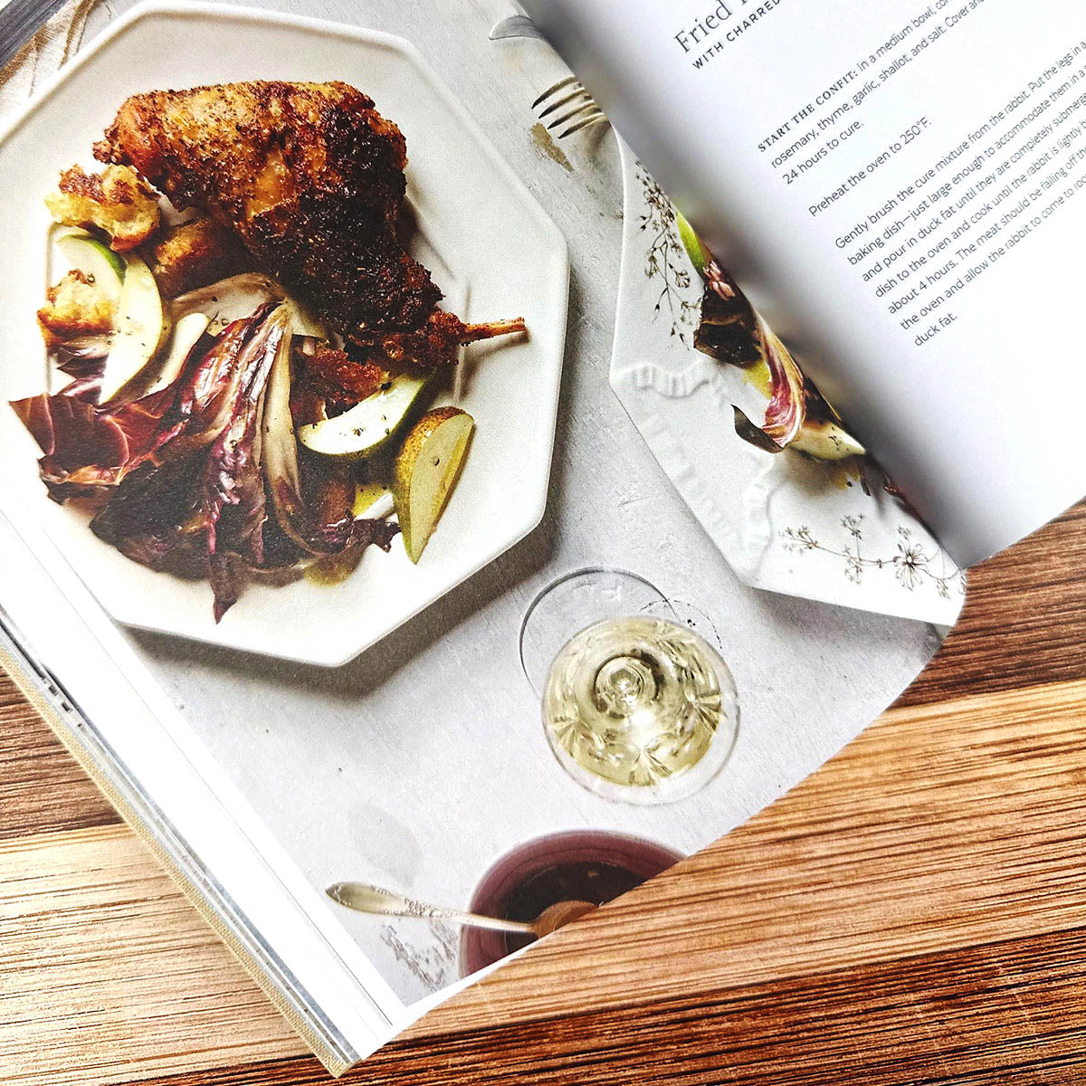 Fried dish with vegetables on a plate, glass of white wine, and book open to a recipe page.