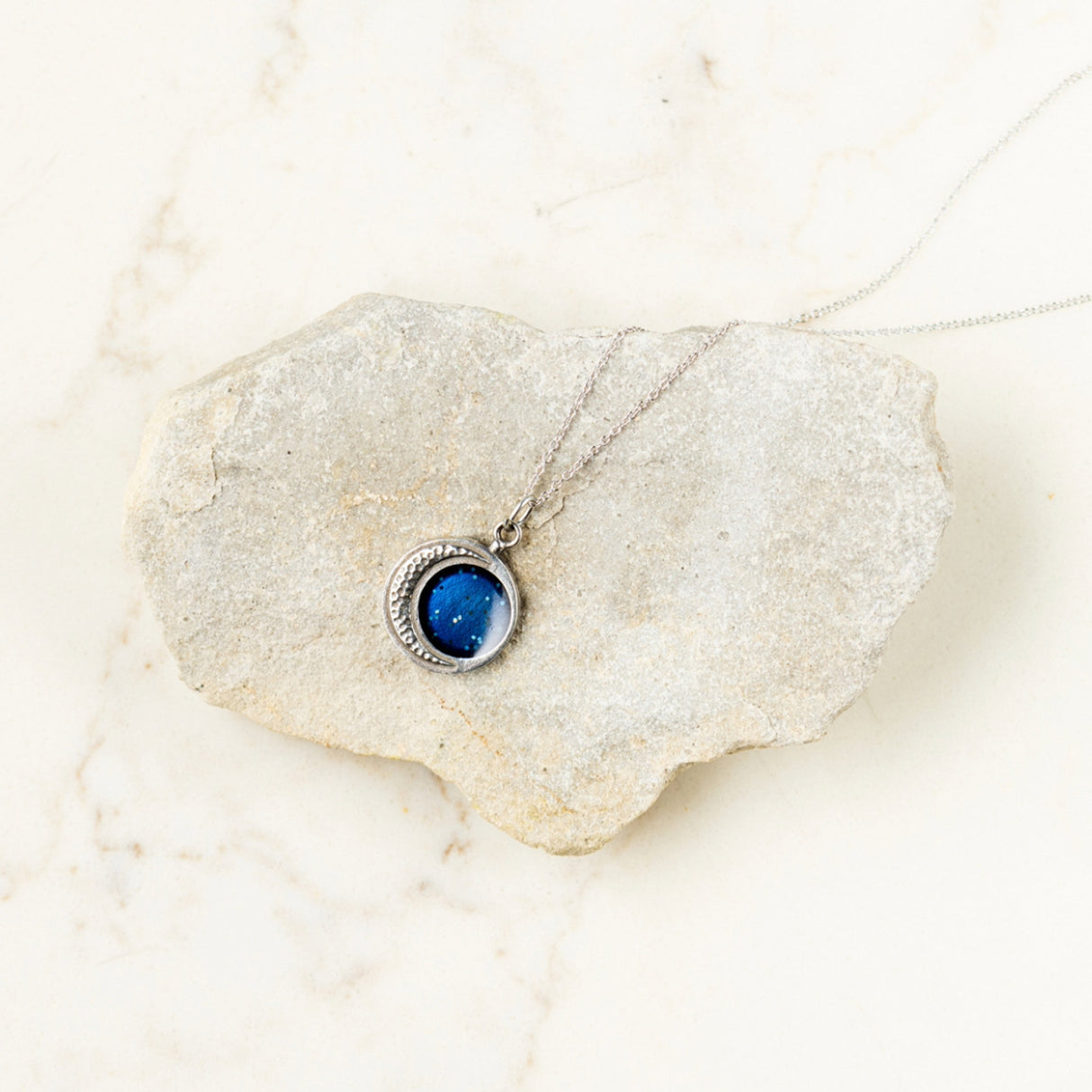 the crescent moon necklace draped over a pale rock that is resting on a marble countertop showing contrasting colors and textures