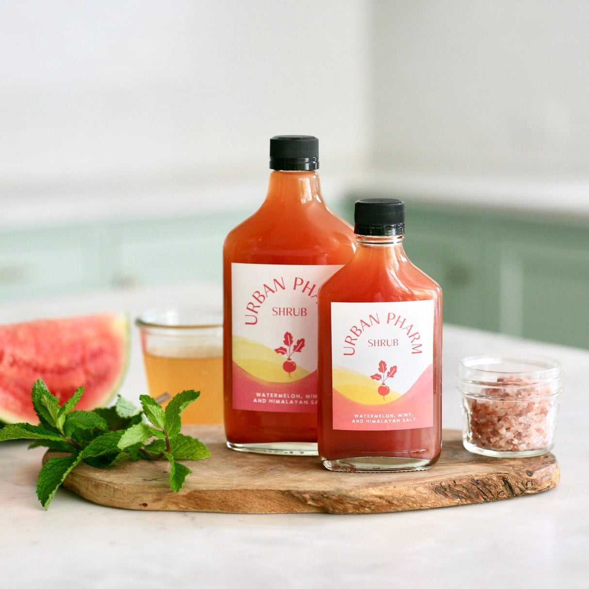 a 200ml and 375ml bottle of watermelon shrub on a wood board surrounded by mint leaves, Himalyan salt, and a slice of watermelon